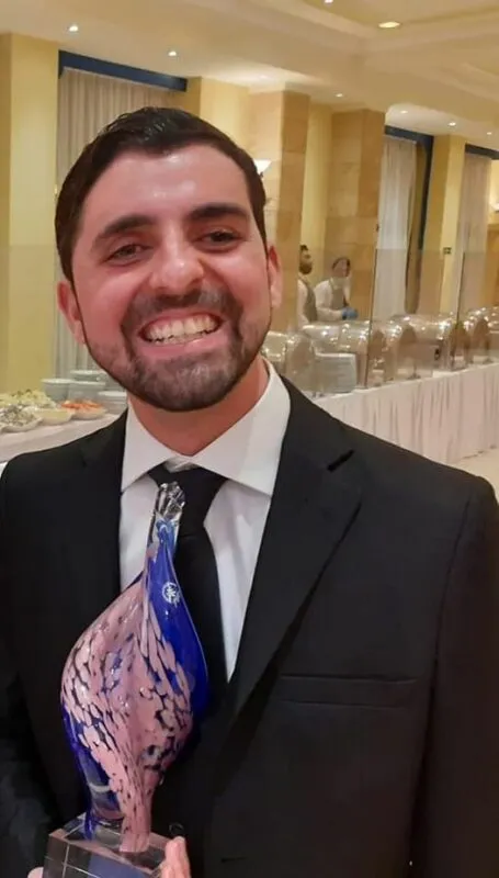 Jacob in a suit, smiling and holding the blue and pink glass Premju Soċjetà Ġusta trophy