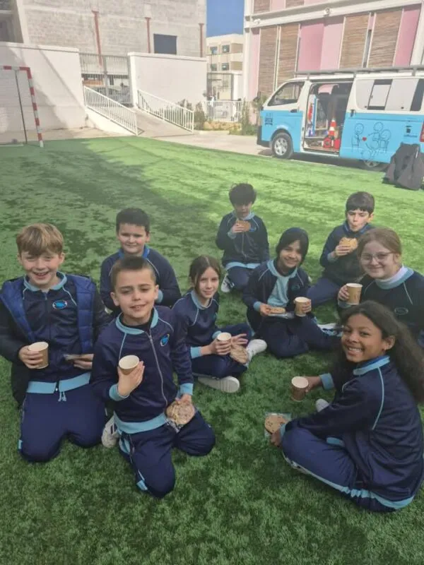 Schoolchildren in navy tracksuits sitting on a sports pitch with hot drinks and cookies, the Jacob's Brew van parked behind them